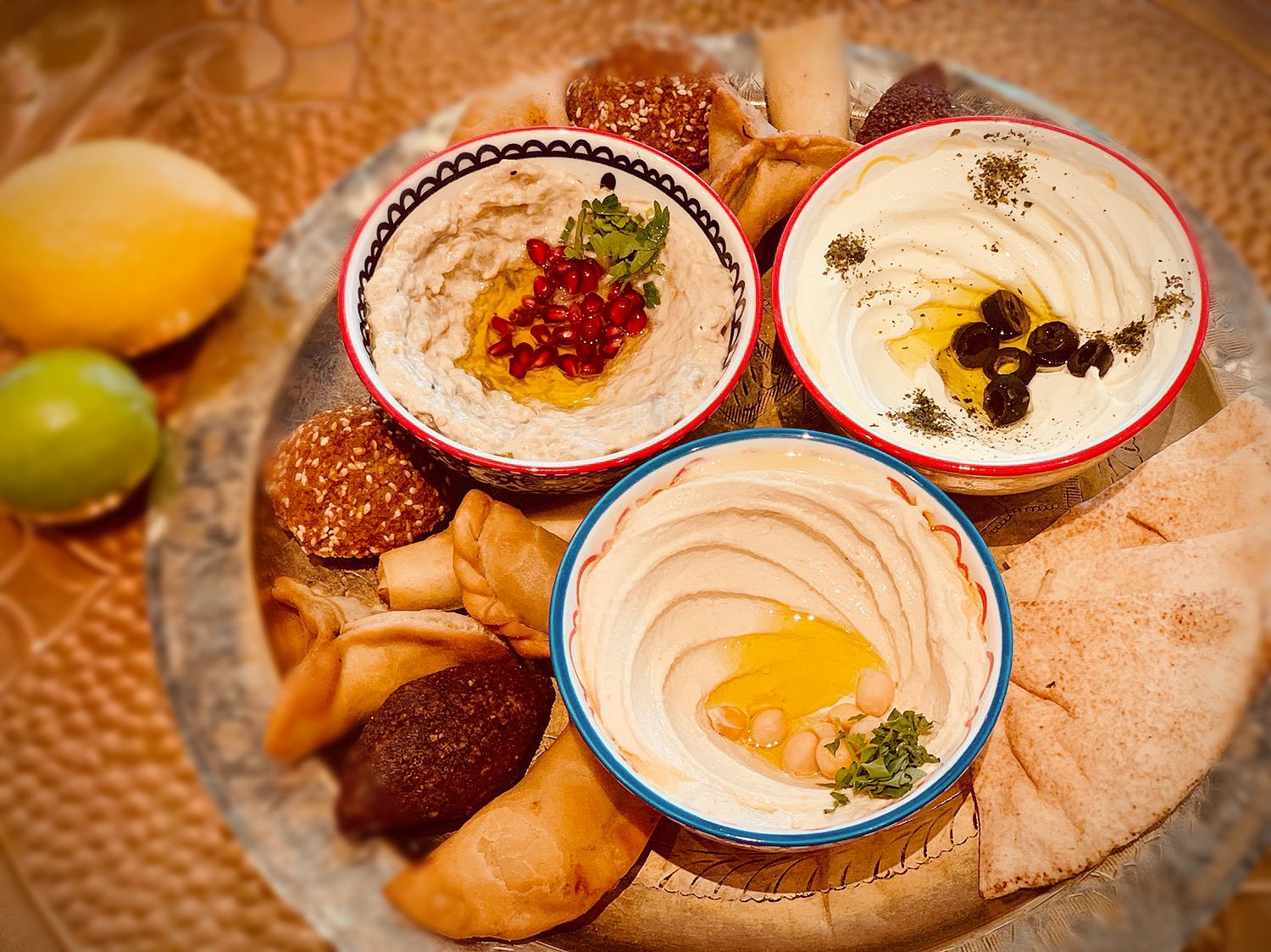 Three bowls of houmous with falafel, kebbeh and pastries on a brass tray